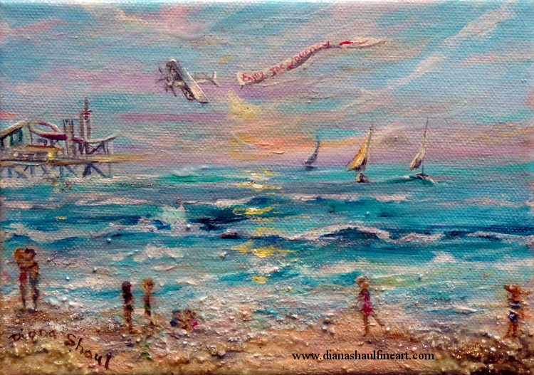 Beach scene with people along shore, a pier in the distance, sailboats on the water and a plane pulling a banner in the sky.