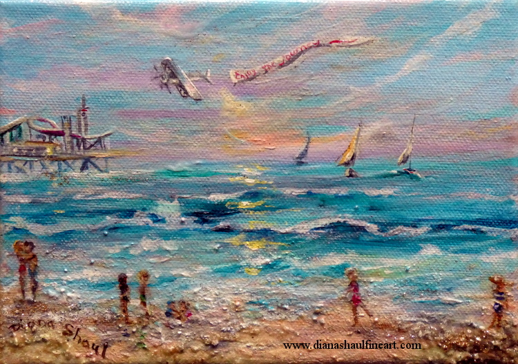 Beach scene with people along shore, a pier in the distance, sailboats on the water and a plane pulling a banner in the sky.